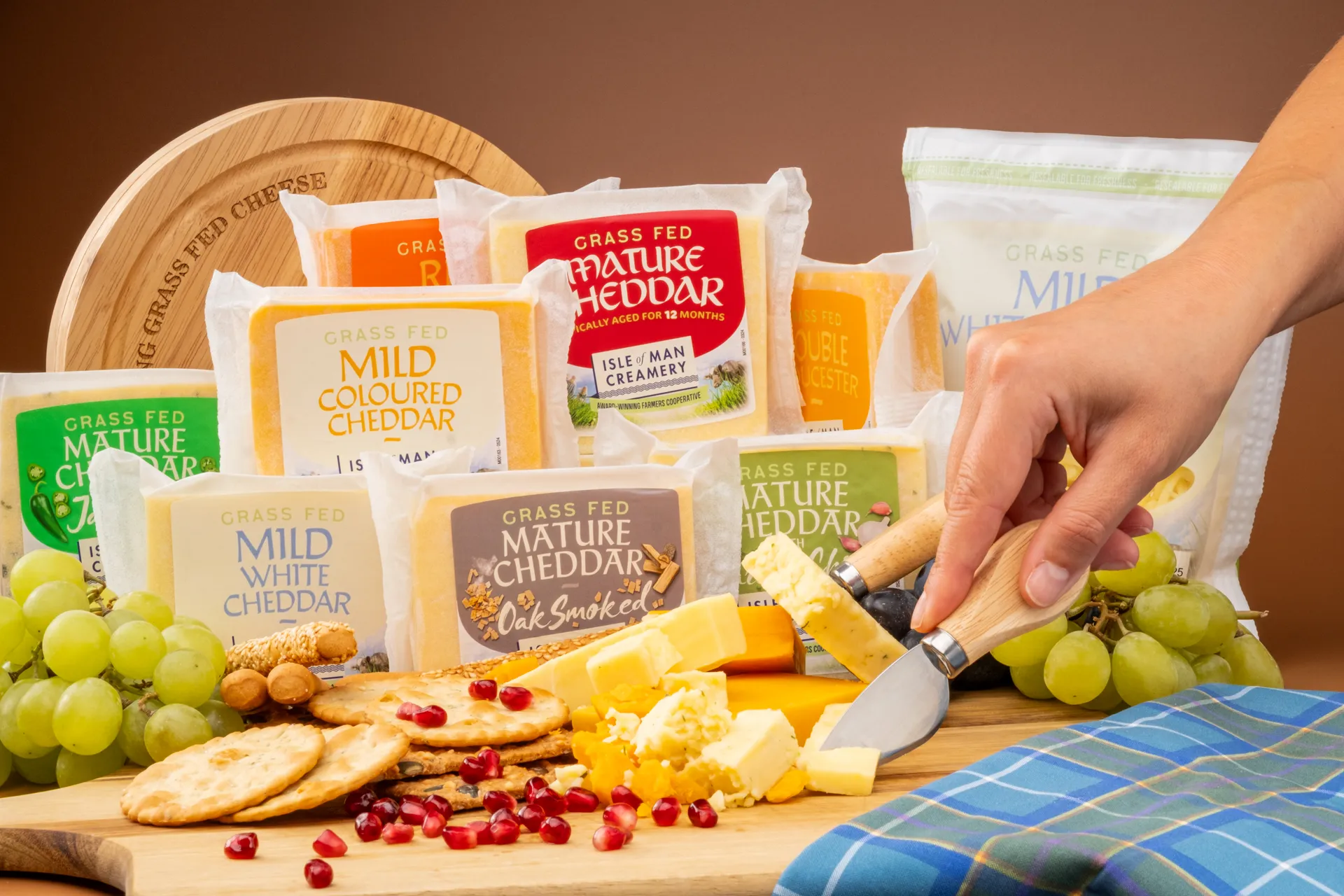 Product photography shot by DotPerformance for the Isle of Man Creamery Doorstep Delivery campaign, showing a range of Isle of Man Creamery grass-fed cheeses including Mature Cheddar, Mild Coloured Cheddar, Oak Smoked Mature Cheddar, and Mild White Cheddar, arranged on a wooden board with crackers, grapes, and pomegranate seeds